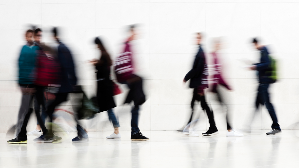 Blurry motion of diverse people walking indoors against a white wall background