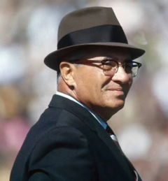 Vince Lombardi in brown fedora black ribbon glasses suit tie smiling on stadium field background